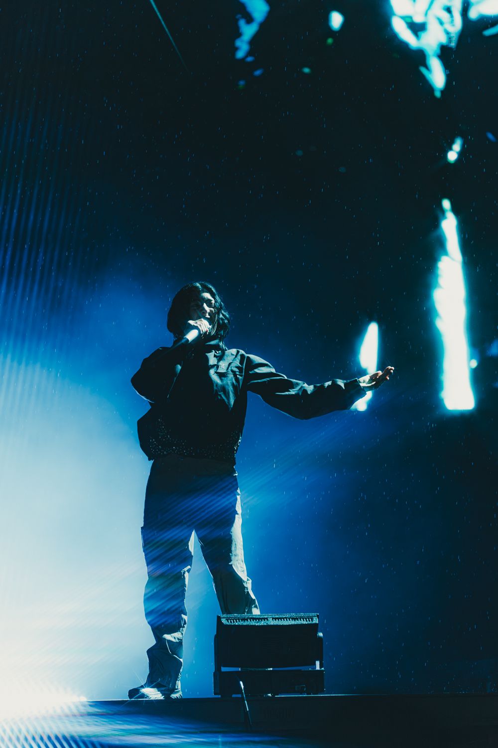 A performer stands on a dark stage holding a microphone, bathed in dramatic blue lighting during a live concert.