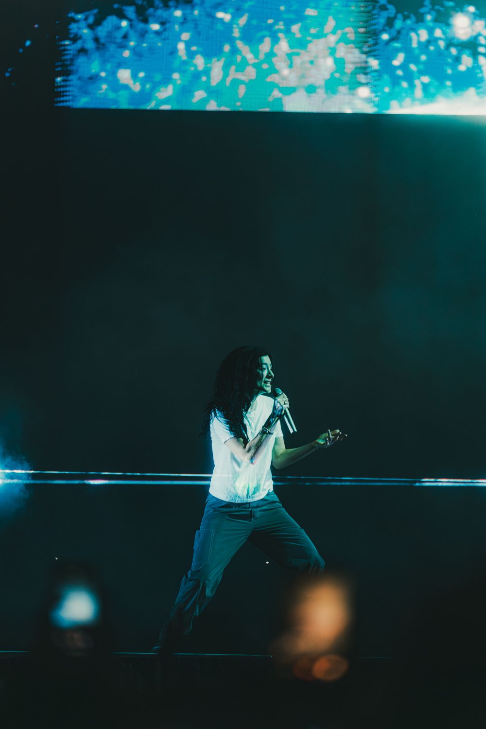 A performer sings on a dimly lit stage with blue lighting and a large screen backdrop during a live concert.