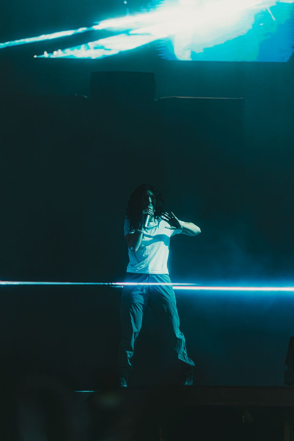 Performer on stage bathed in teal neon light with dramatic fog and bright beam effects during live concert.