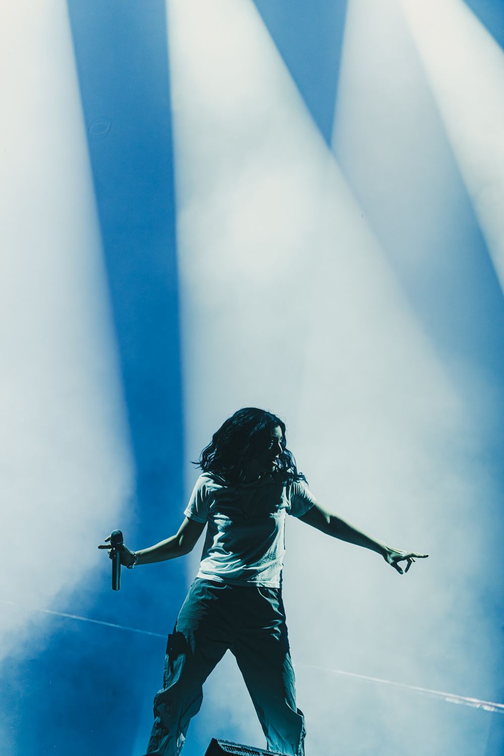 Performer with dreadlocks holds microphone on stage, arms outstretched, bathed in dramatic blue spotlights.