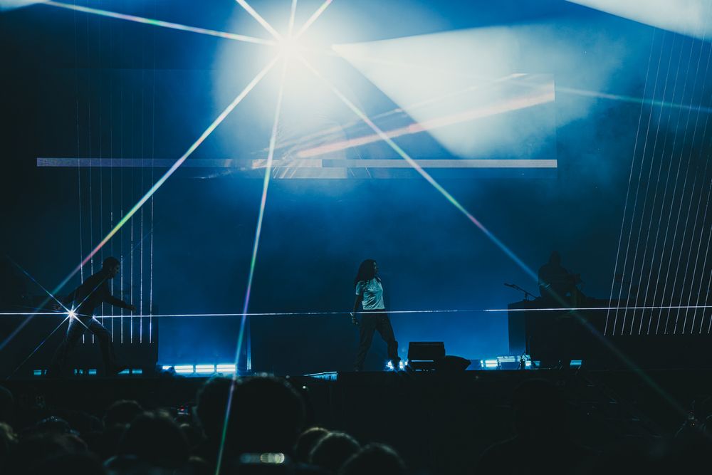DJ performing on stage with dramatic blue laser light show and silhouetted crowd at a live concert event.
