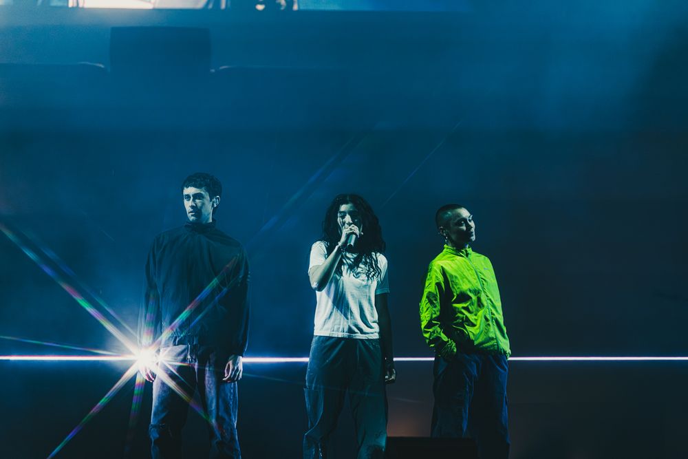 Three performers stand on a dramatic blue-lit stage with laser beams during a live concert performance.