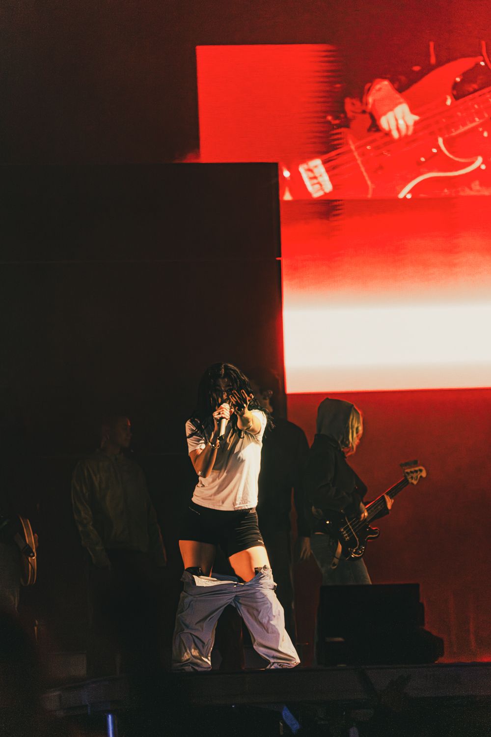 Female singer performing on stage with guitarist and red backdrop lighting at live concert.
