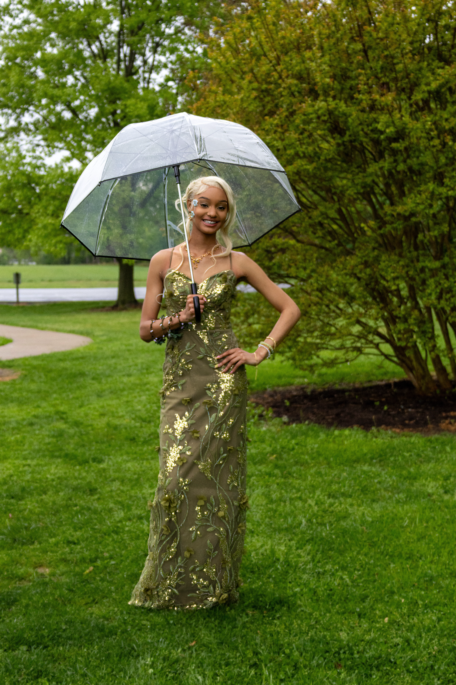 Young woman in gold sequin gown holding clear umbrella, posing outdoors on green lawn near trees.