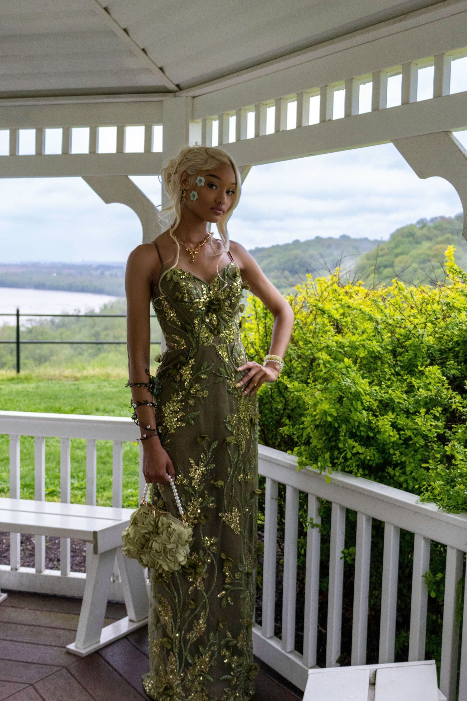 Young woman in elegant olive green floral embroidered gown posing on white gazebo porch with scenic green landscape.