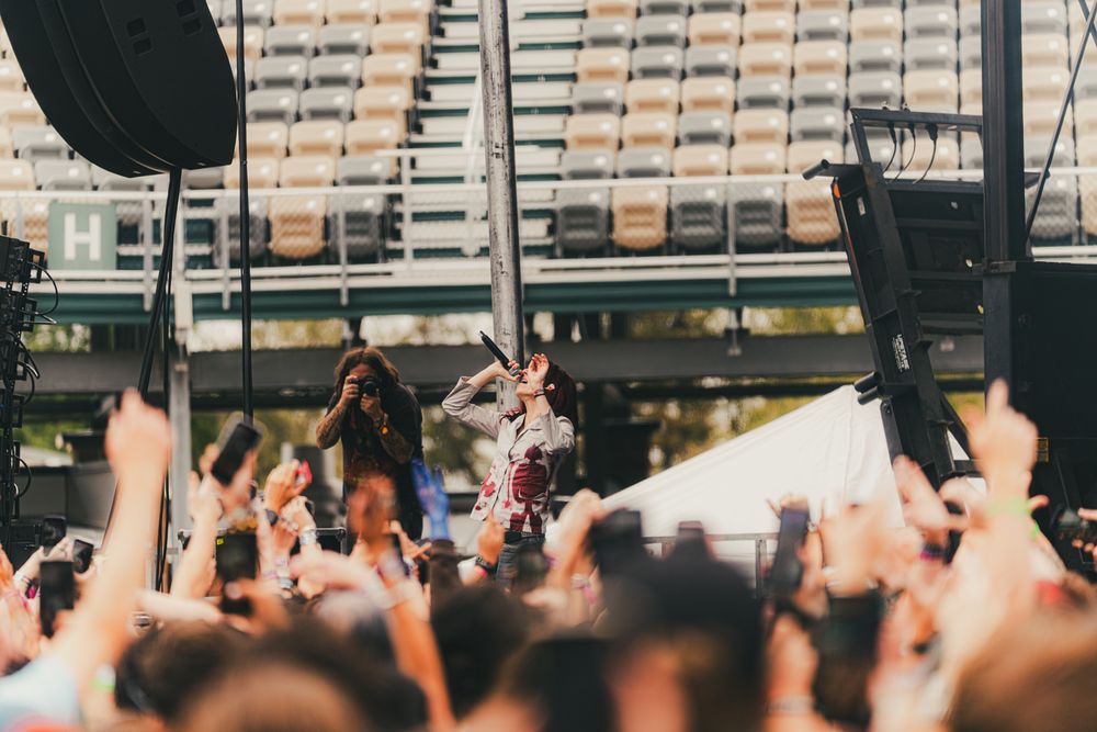 Energetic performer on outdoor festival stage singing into microphone as crowd reaches up with hands.
