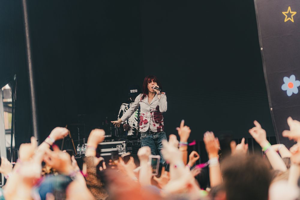 Female singer performing on stage at night with crowd raising hands toward colorful decorated backdrop.