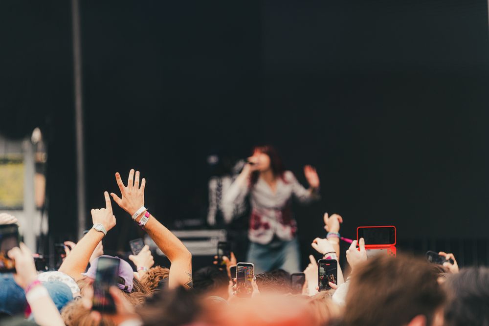 Concert crowd with raised hands filming performer on outdoor festival stage with smartphones.