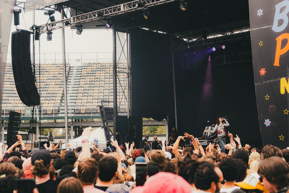 Crowd of fans at an outdoor music festival concert stage with performers and large speakers under an overcast sky.