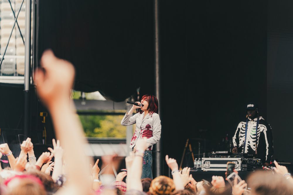Singer performs on outdoor stage before large crowd with skeleton-costumed musician and raised hands in foreground.