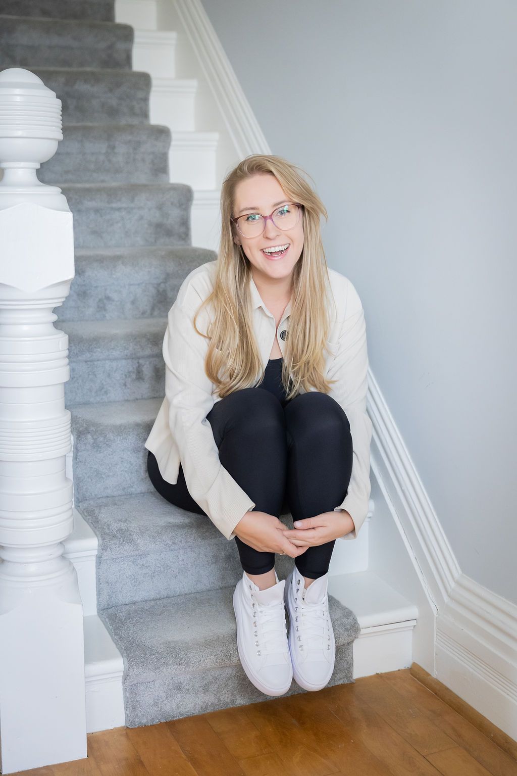 Smiling blonde woman with glasses sitting on carpeted stairs in white sneakers and black leggings.