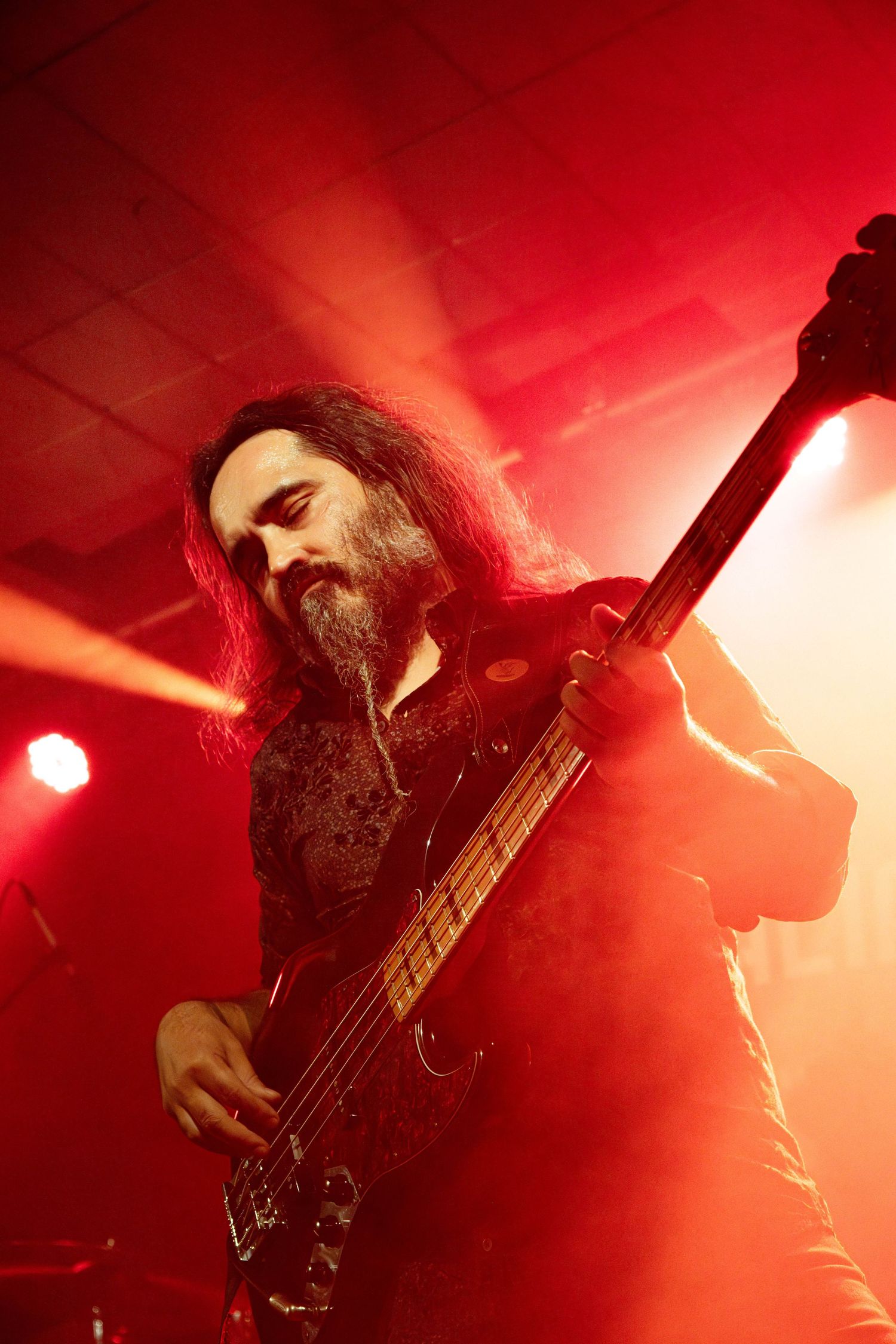 Bearded male guitarist performing on stage under dramatic red lighting in a dark rock concert venue.