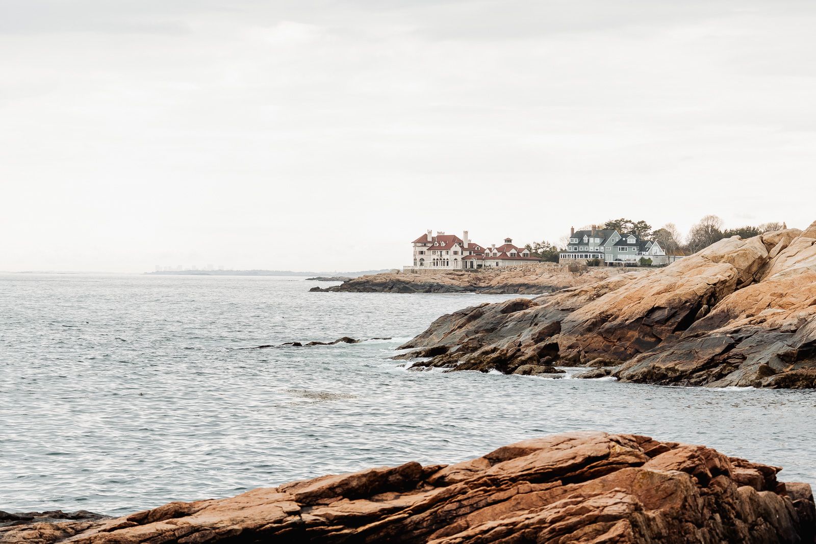 Rocky coastline with historic homes perched on rugged cliffs overlooking calm gray ocean waters under an overcast sky.