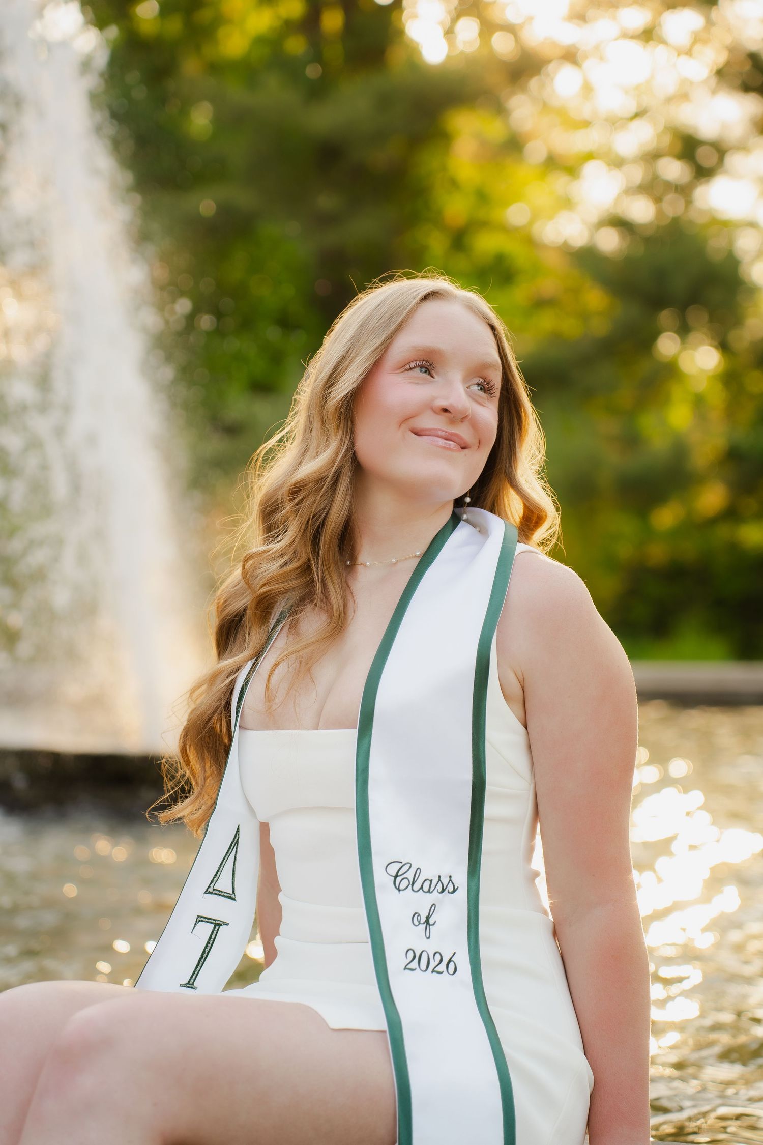Smiling college student wearing a Delta Tau Class of 2026 graduation sash, seated near a fountain outdoors.