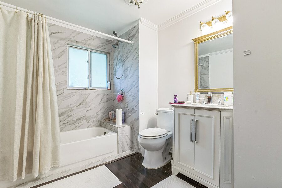 Bright bathroom with marble-tiled tub surround, white vanity with gold mirror, hardwood floors, and a window above the tub.