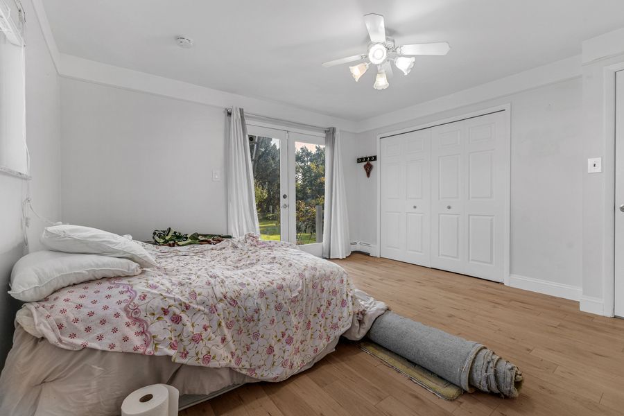 Bright bedroom with hardwood floors, floral bedding, ceiling fan, large window with garden view, and white closet doors.