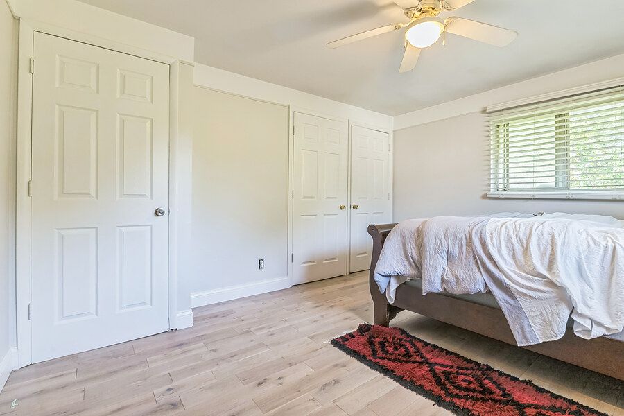 Bright bedroom with white walls, hardwood floors, ceiling fan, two closet doors, and a bed with white bedding and red rug.
