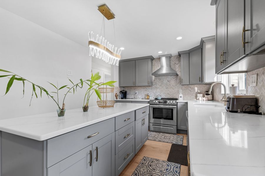 Modern kitchen with gray shaker cabinets, white quartz countertops, stainless steel range, and gold pendant lighting.