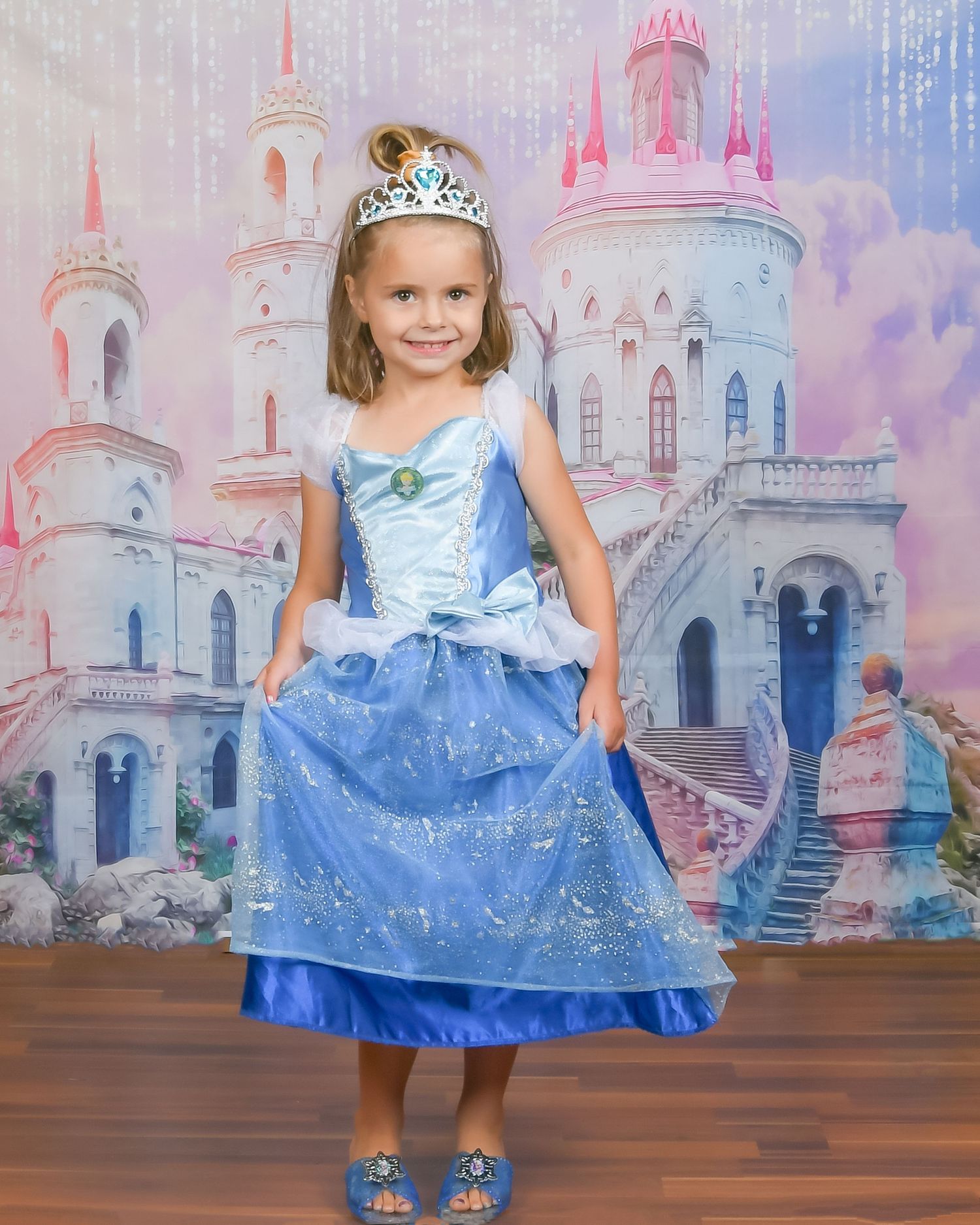 Young girl in blue Cinderella princess dress and tiara poses in front of fairy tale castle backdrop.