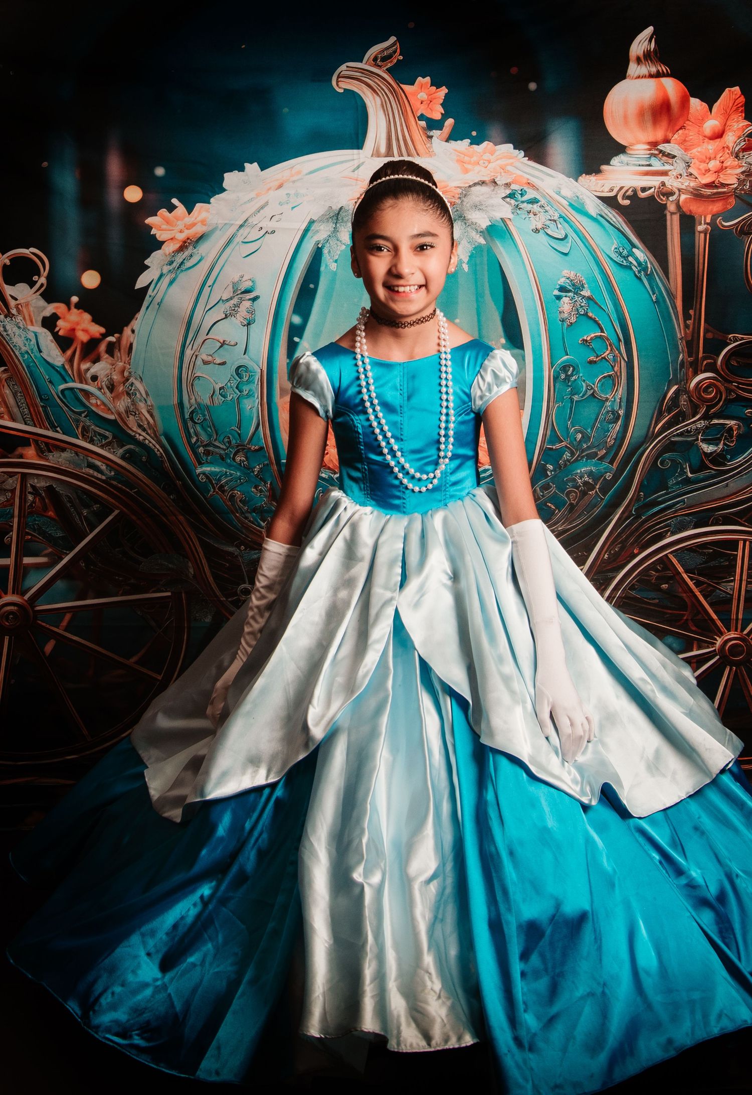 Young girl in Cinderella blue ball gown poses before enchanted pumpkin carriage backdrop.