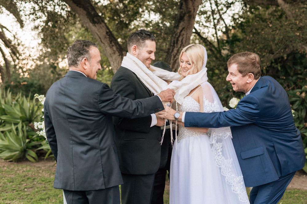 Bride and grooms fathers wrap couple in ceremonial shawls