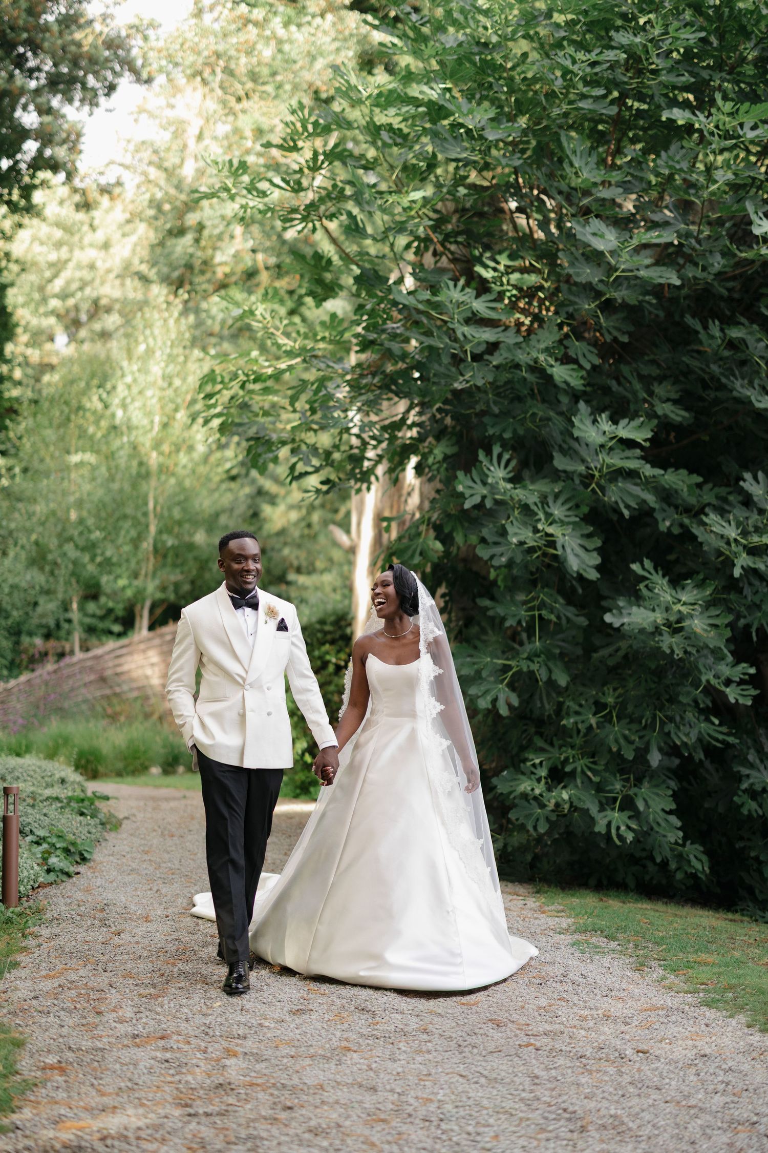 A couple in wedding attire walks along a tree-lined gravel path during their outdoor garden ceremony.