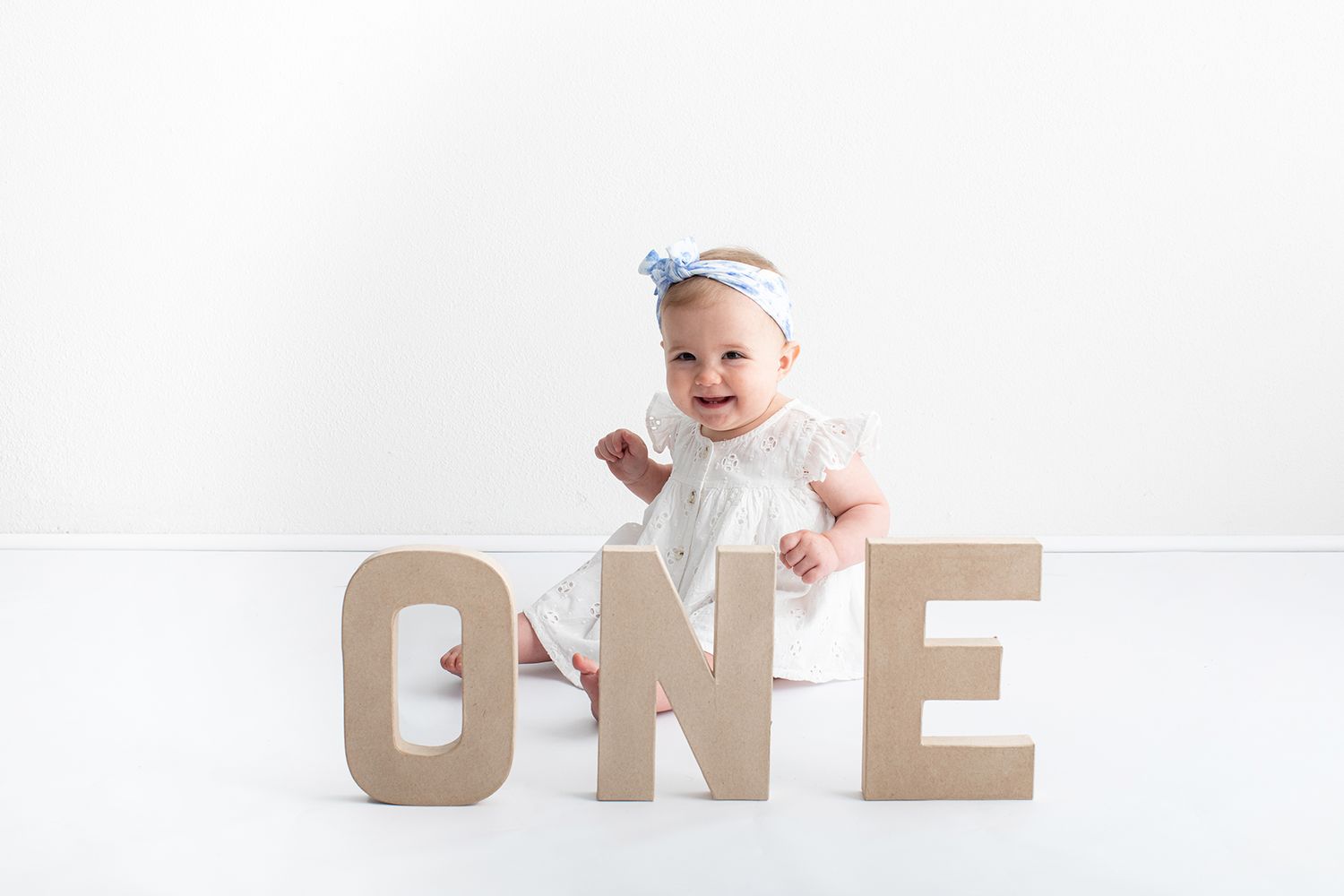Baby girl in white dress celebrates her first birthday sitting behind large cardboard ONE letters with a bow headband.