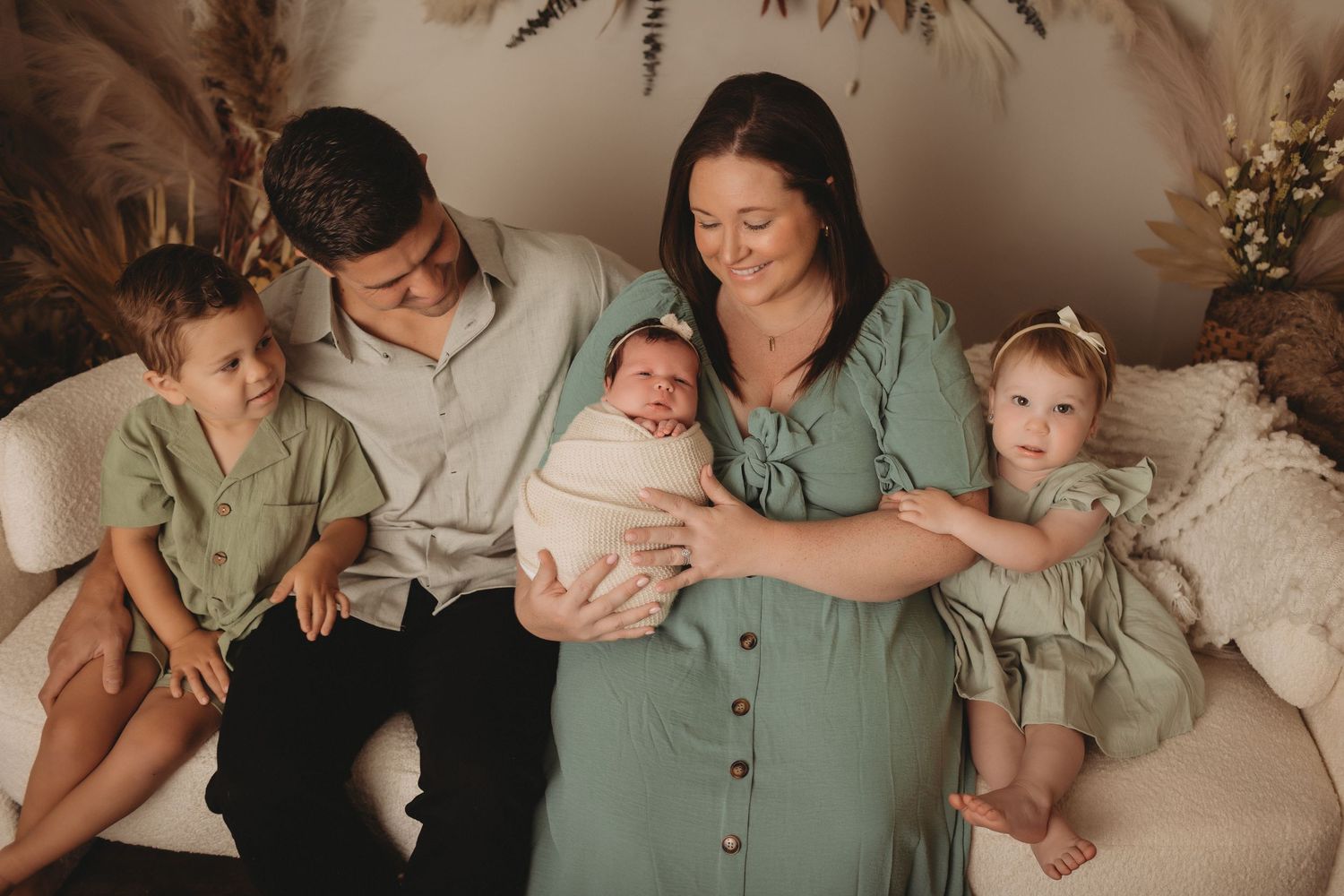 Family of five poses on a couch, parents holding newborn baby, with two young children in matching green outfits.