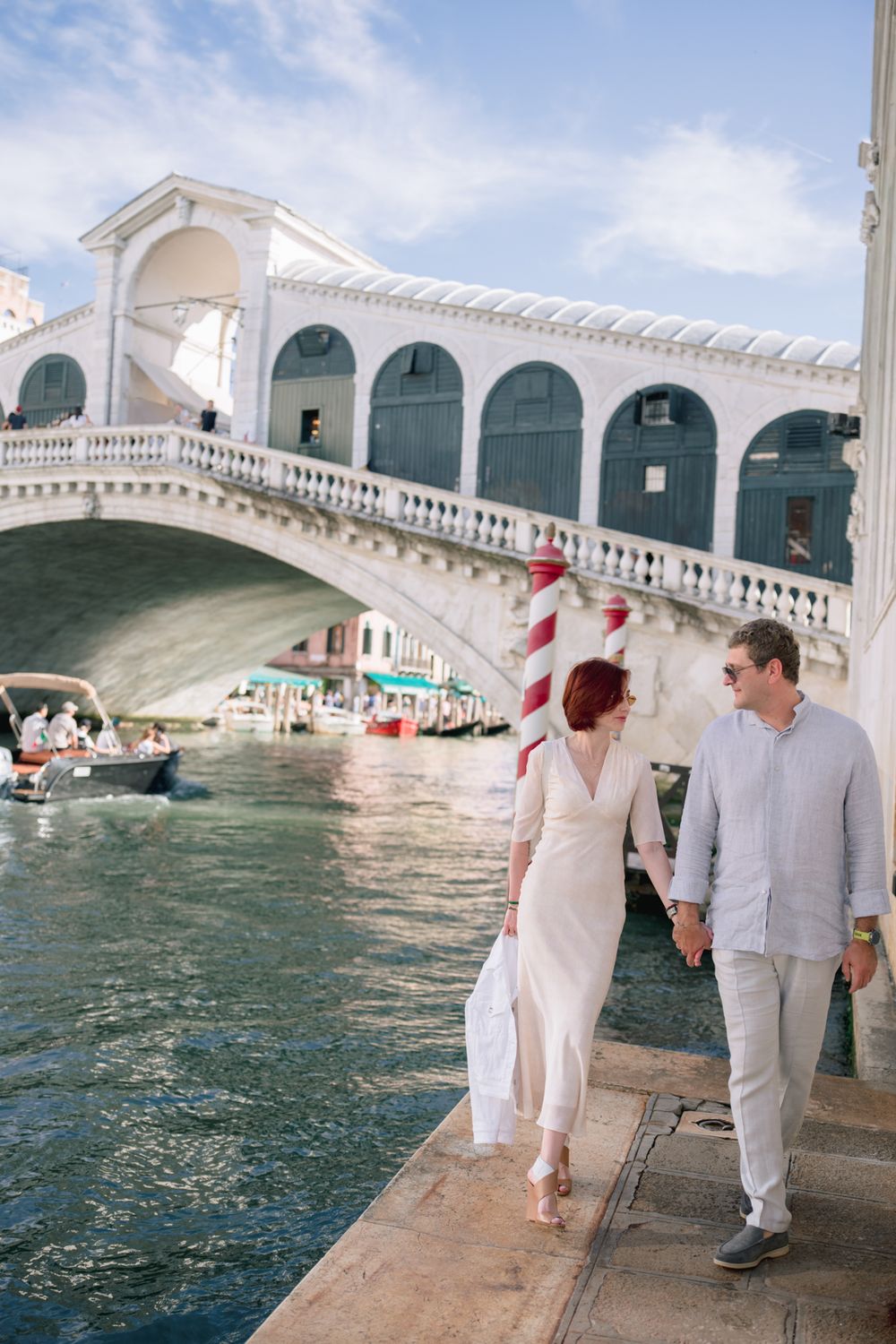 Couple holding hands walking along Grand Canal near Rialto Bridge in Venice, Italy on a sunny day.