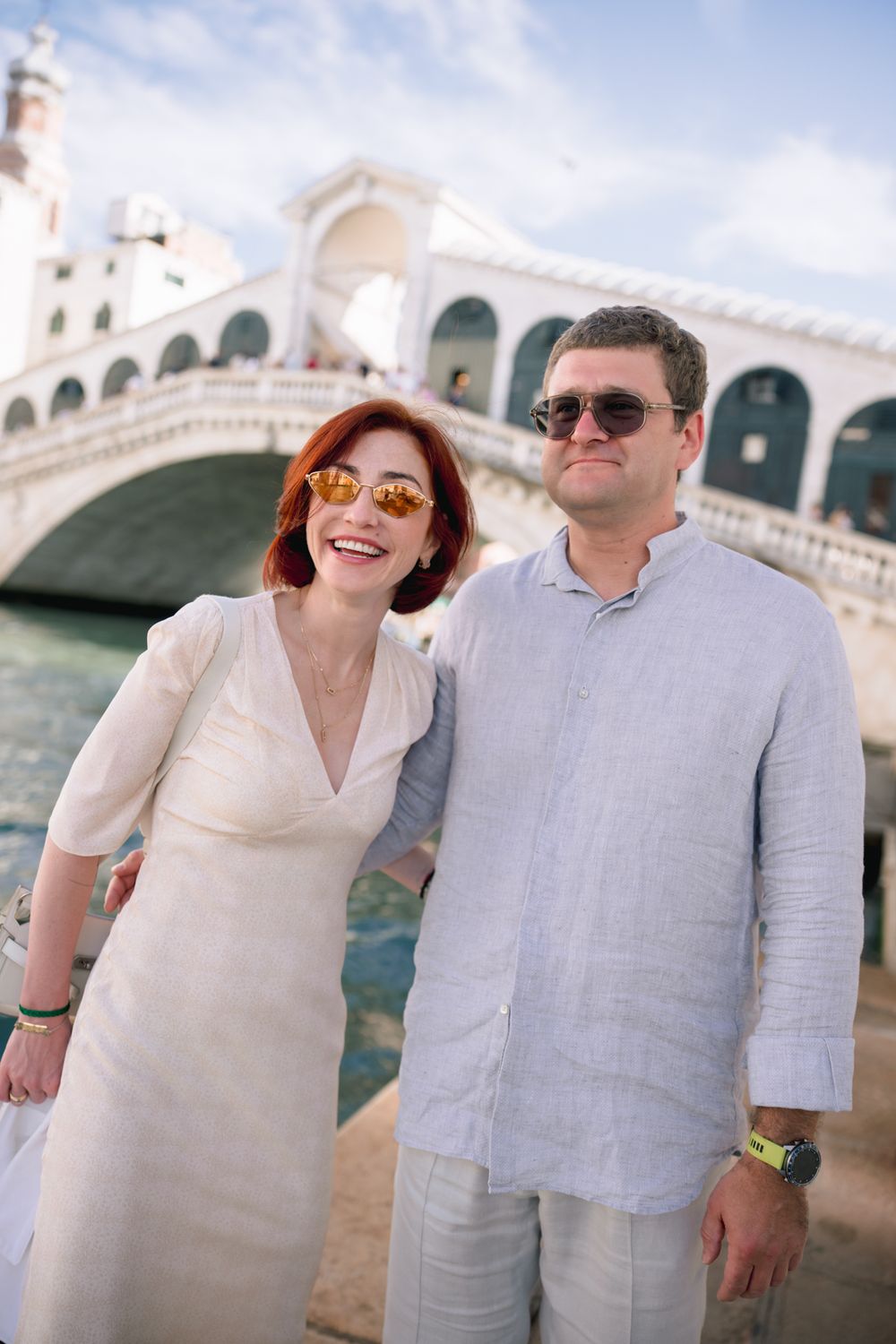 Smiling couple in sunglasses posing in front of the Rialto Bridge in Venice, Italy on a sunny day.