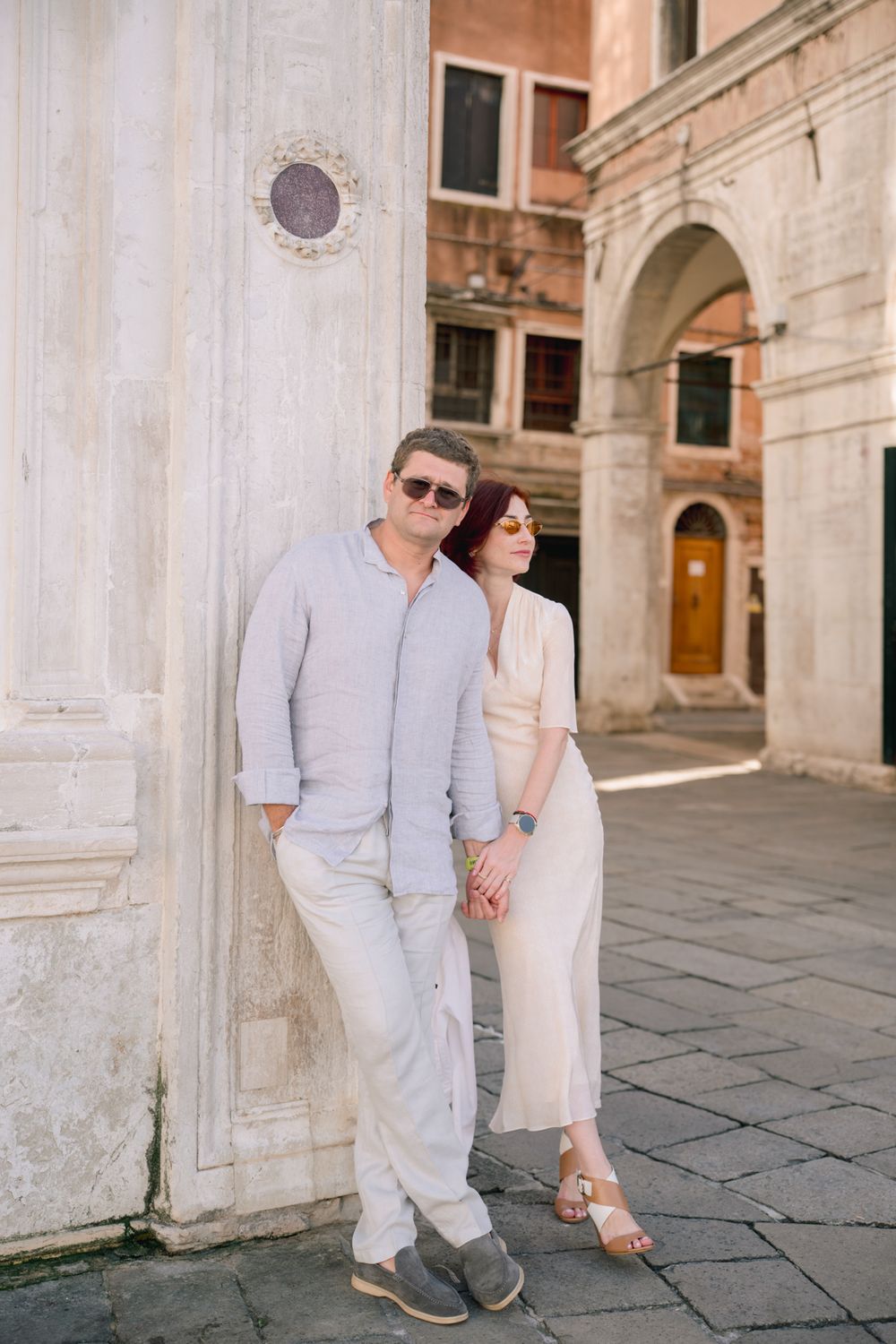 Couple in light summer outfits leaning against a white marble wall in a sunny Venice piazza, holding hands.
