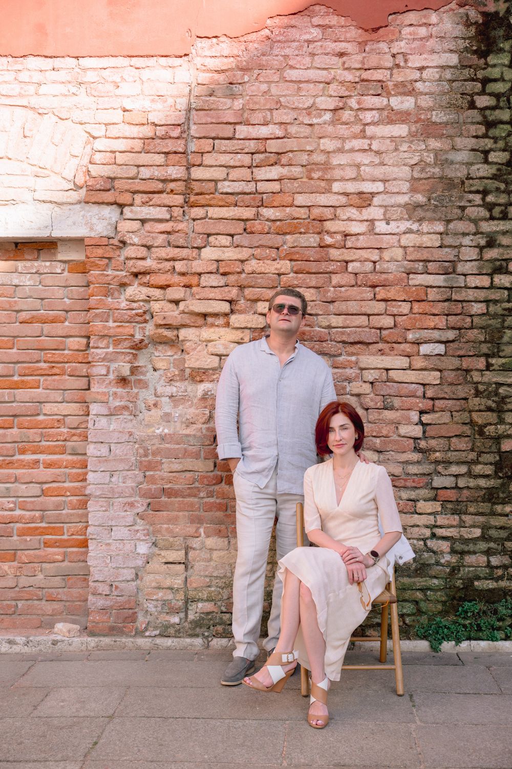A couple poses against a rustic brick wall, woman seated in wooden chair, both wearing light-colored outfits on stone pavement.
