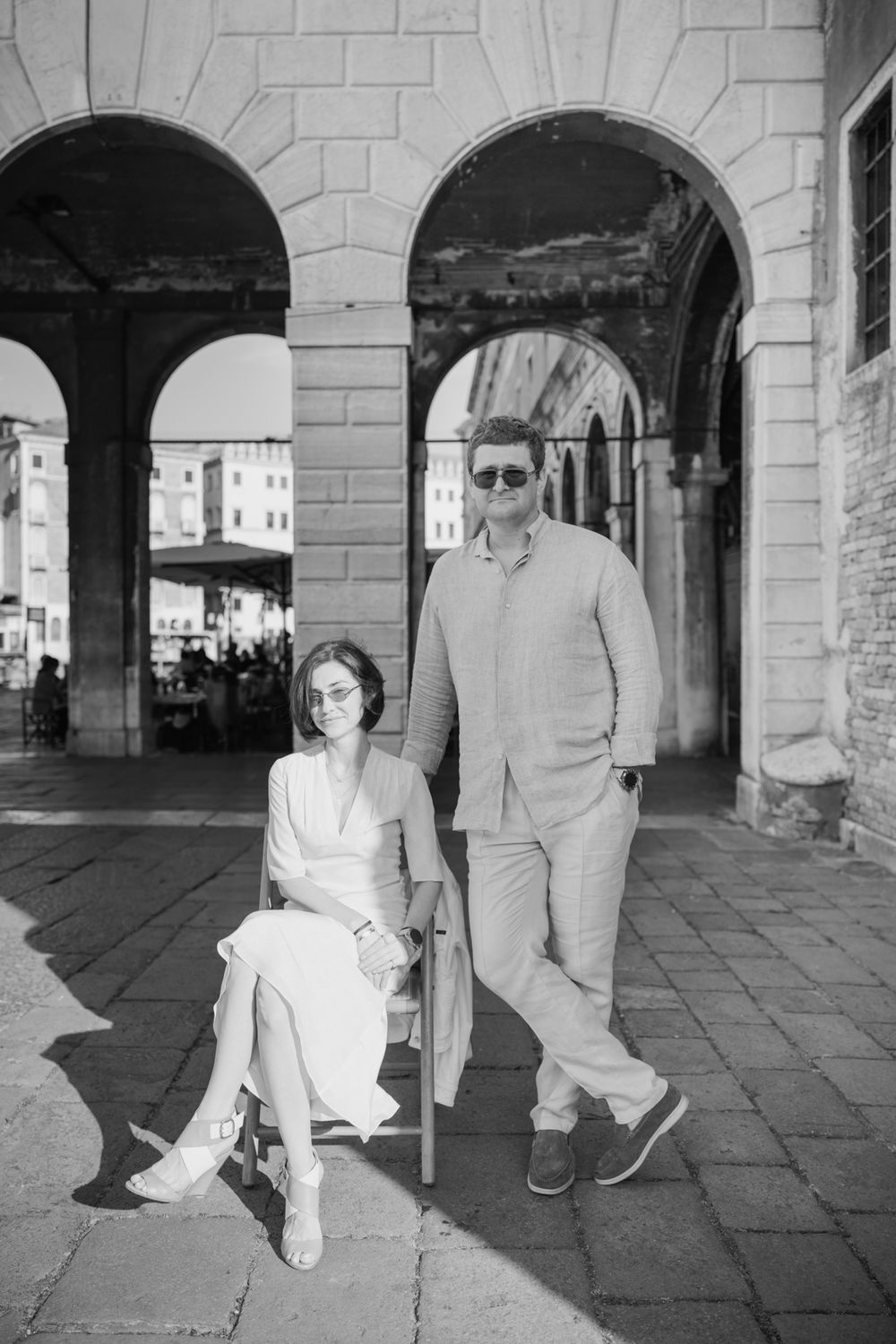 Black and white photo of a couple posing under stone arches in Venice, woman seated on chair, man standing beside her.