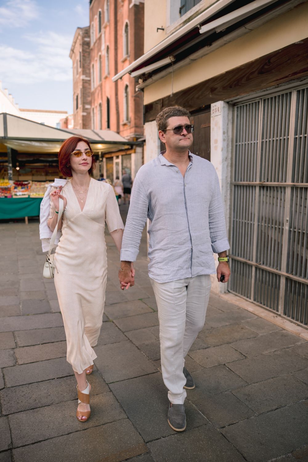 Couple in elegant white outfits holding hands while strolling through a European city street on a sunny day.