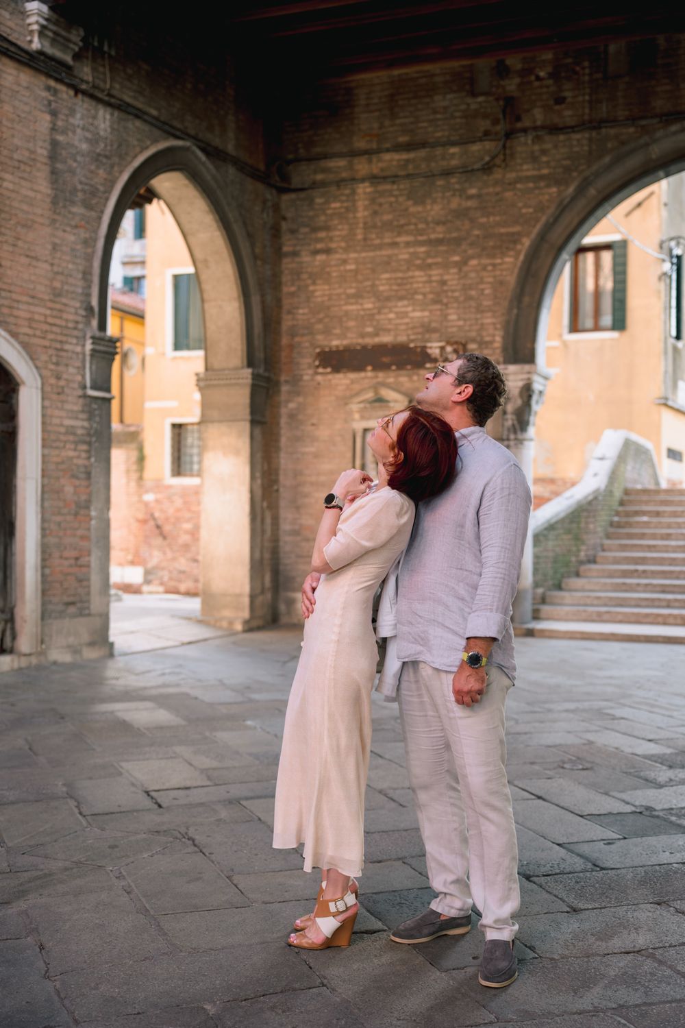 Couple embracing under stone arches in Venice, Italy, with historic brick buildings and a bridge in the background.