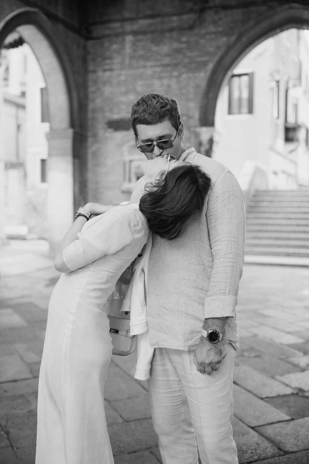 Black and white photo of a couple embracing tenderly under a stone arch in a European city square.