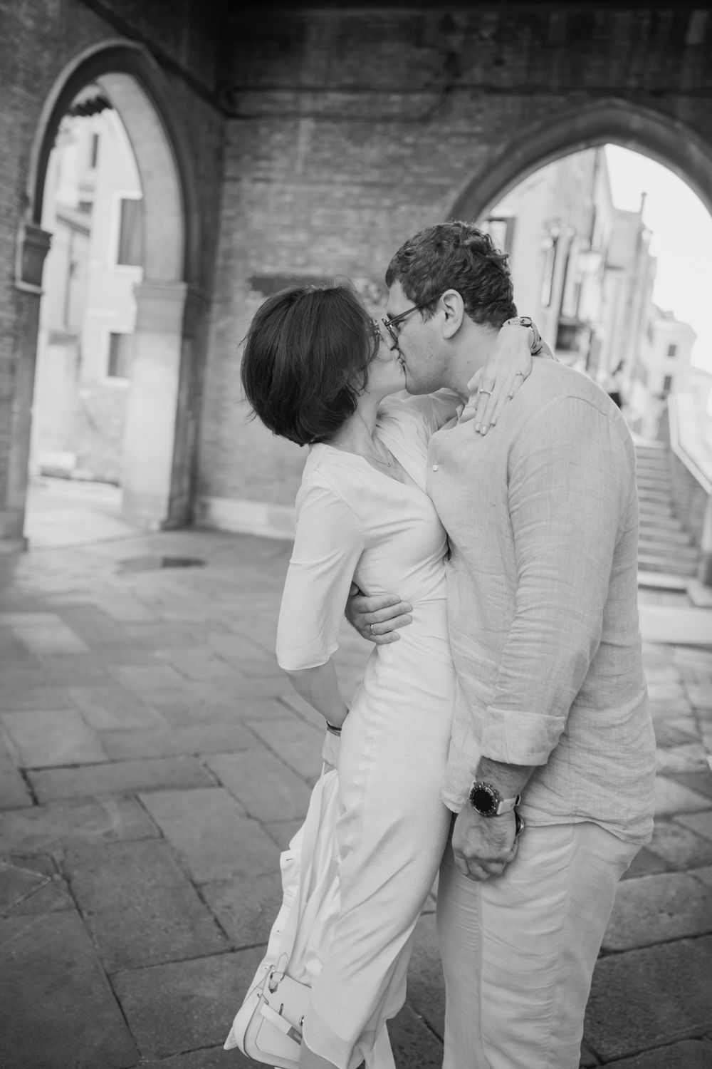 Black and white photo of a couple sharing a romantic kiss under stone arches in a European street setting.