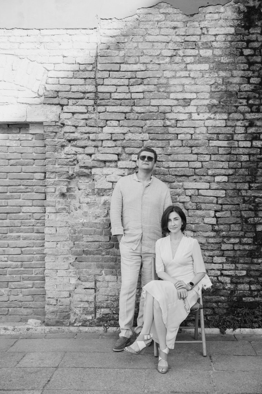 Black and white photo of a couple posing against a rustic brick wall, woman seated on chair, man standing beside her.