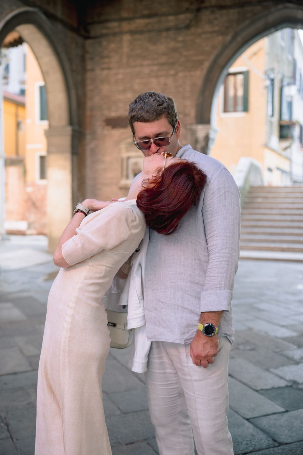 Couple embracing in Venice, woman in white dress with red hair, man in linen suit with sunglasses, stone archway background.