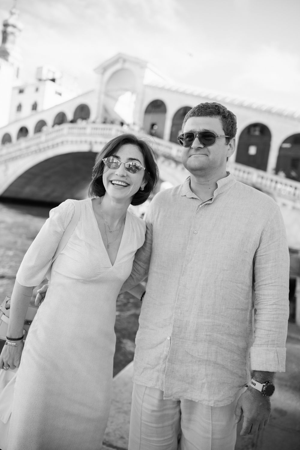 Couple posing in front of the Rialto Bridge in Venice, wearing sunglasses and light outfits, black and white photo.