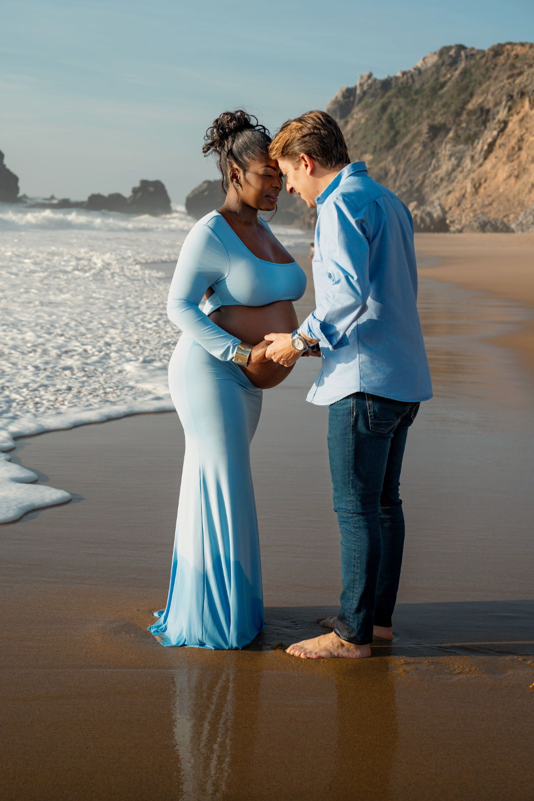 Pregnant couple in blue outfits holding hands on a sandy beach with ocean waves and rocky cliffs.