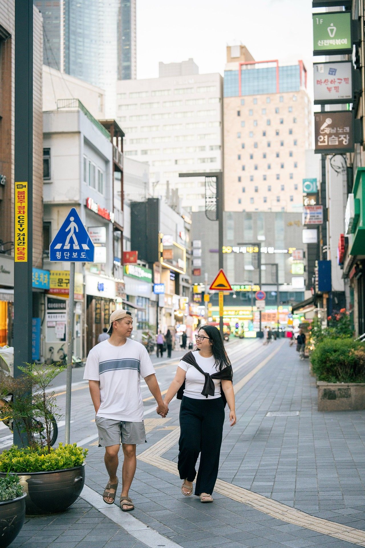 Couple holding hands and walking together on a busy Korean street lined with colorful signs and urban buildings.