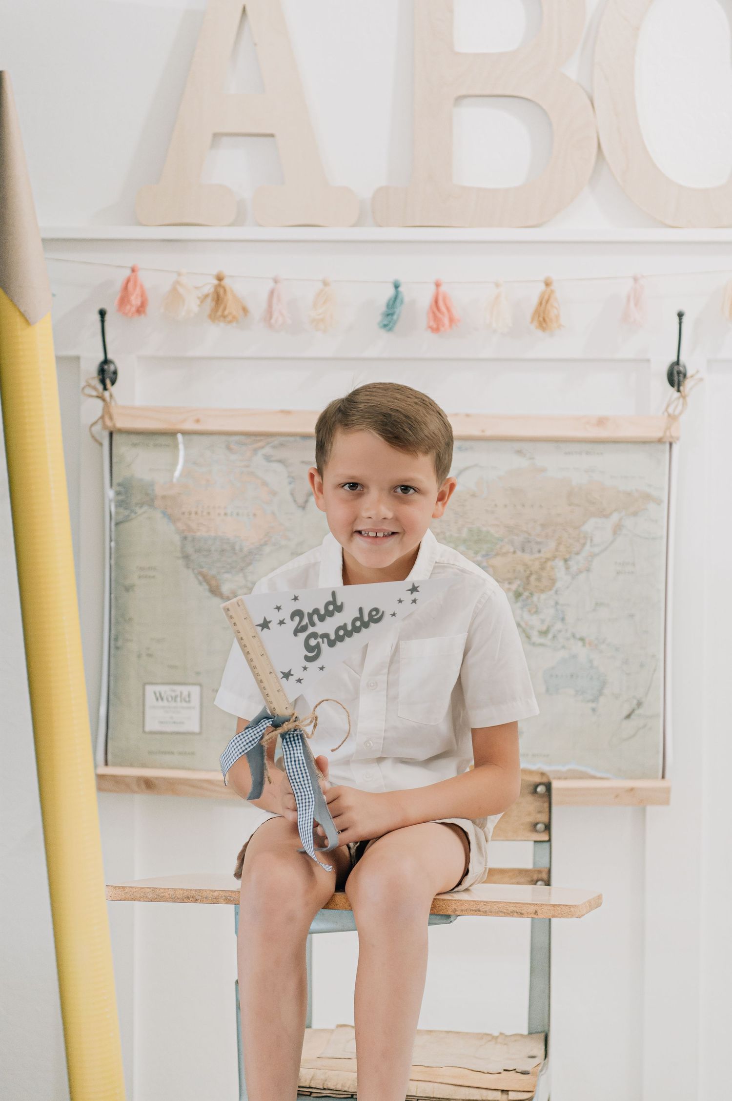 A smiling boy sits holding a 2nd Grade sign, with ABC letters and a world map backdrop. Glendale minis Arizona