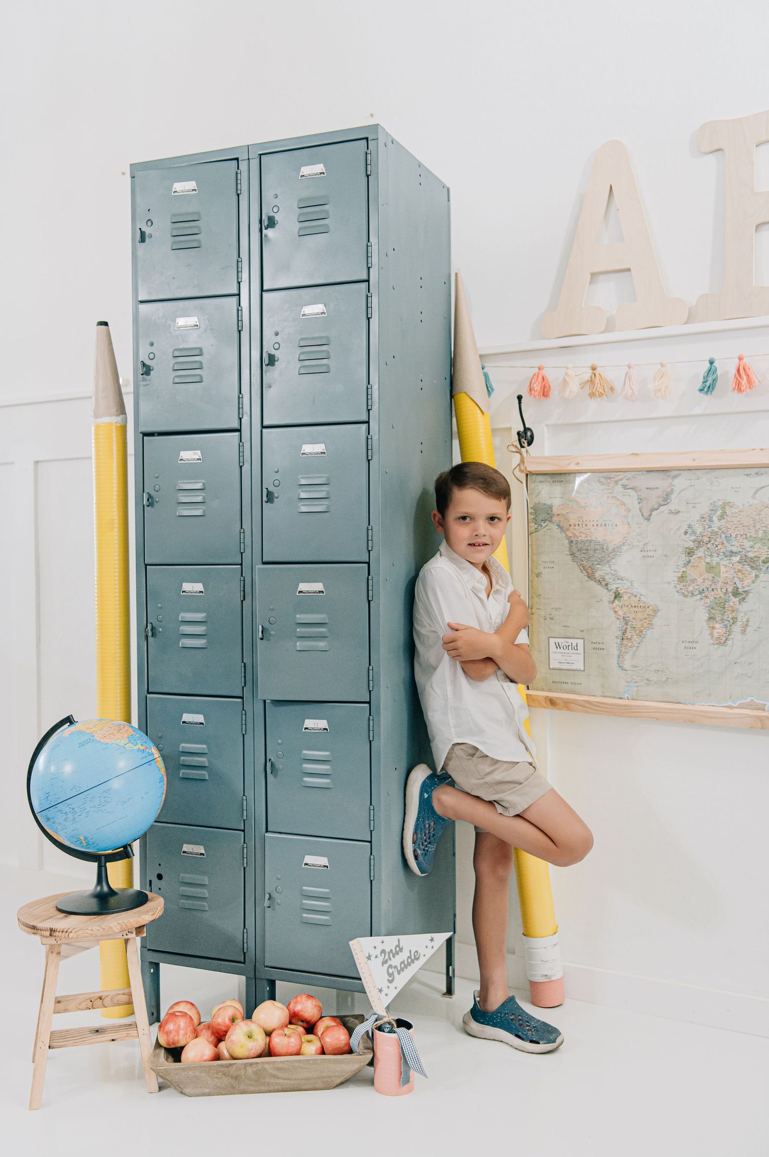 Boy leaning against gray school lockers surrounded by giant pencils, globe, apples, and a world map in a bright classroom. Back to school
