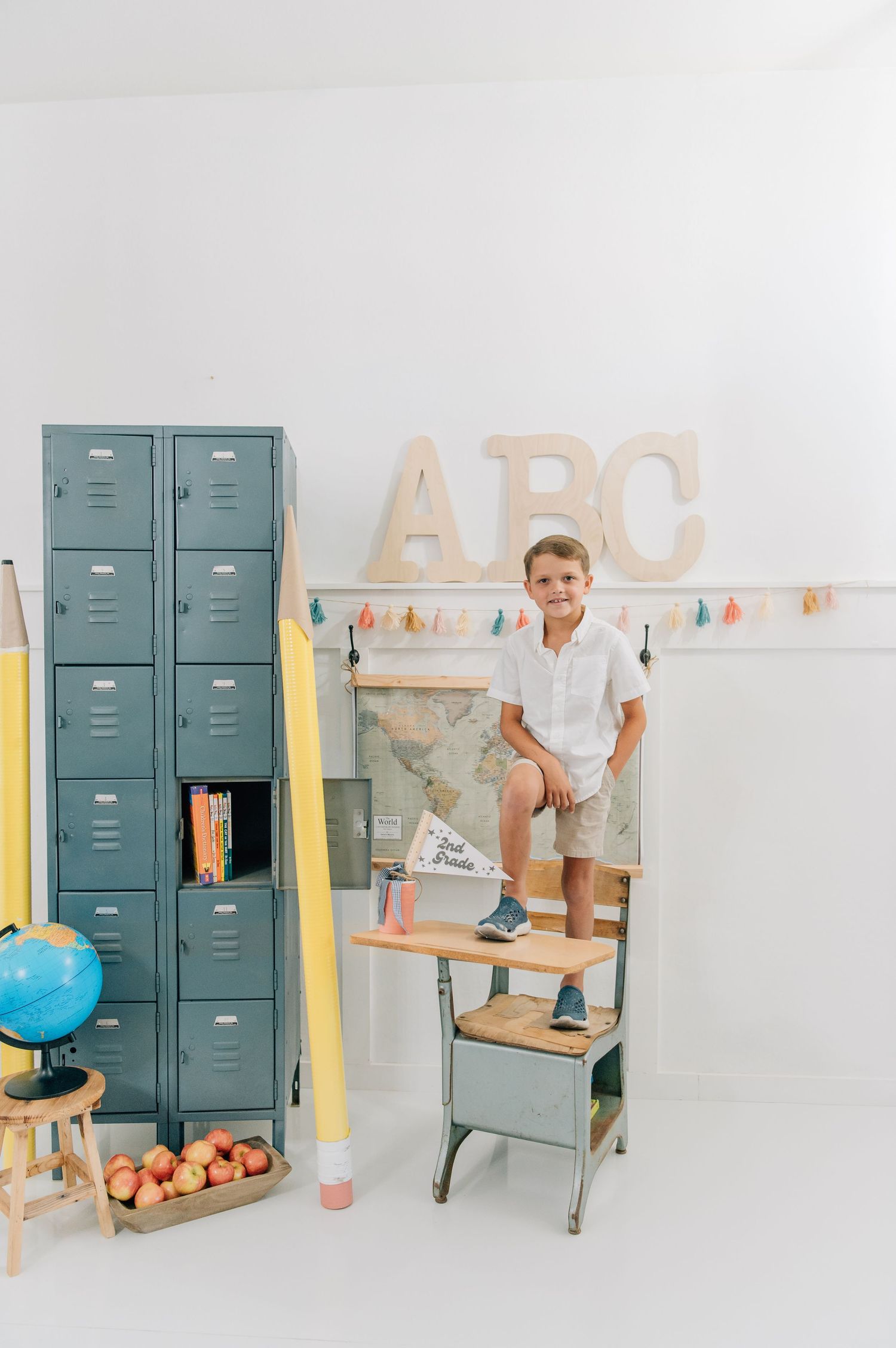 Boy sitting on vintage school desk beside teal lockers with ABC letters and school supplies in bright classroom. Back to school minis