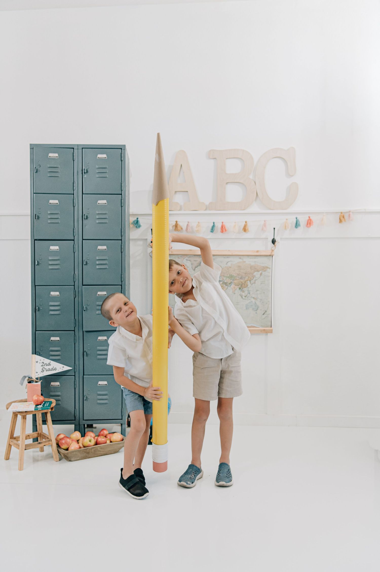 Two children pose with a giant yellow pencil in a bright classroom with teal lockers and ABC letters on the wall. Glendale, Peoria, Arizona