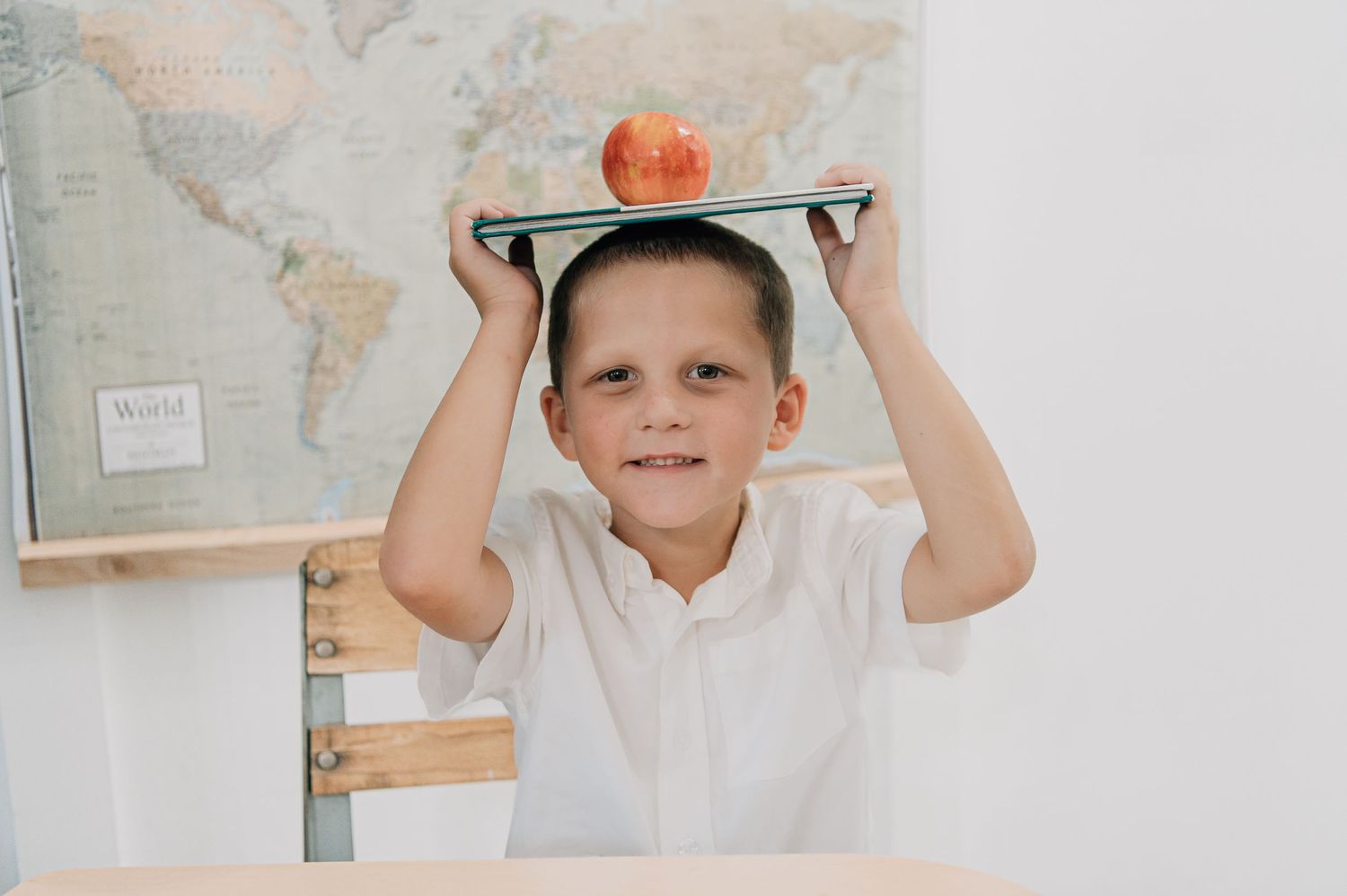 A smiling boy in a white shirt balances a book with an apple on his head in front of a world map.