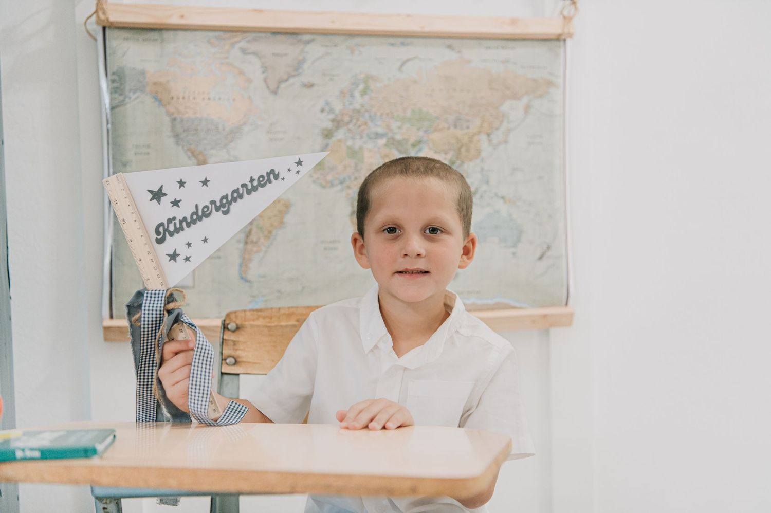 Young child sitting at a school desk holding a Kindergarten pennant flag with a world map in the background. Back to school minis Arizona