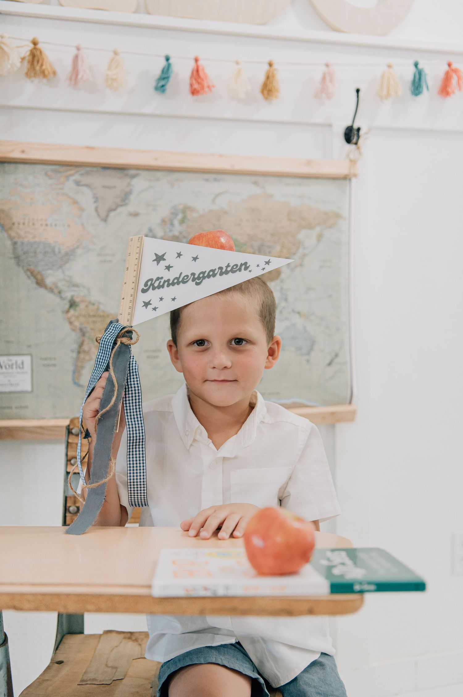 A young boy sits at a desk wearing a Kindergarten pennant hat, with an apple and backpack, ready for his first day of school. Glendale