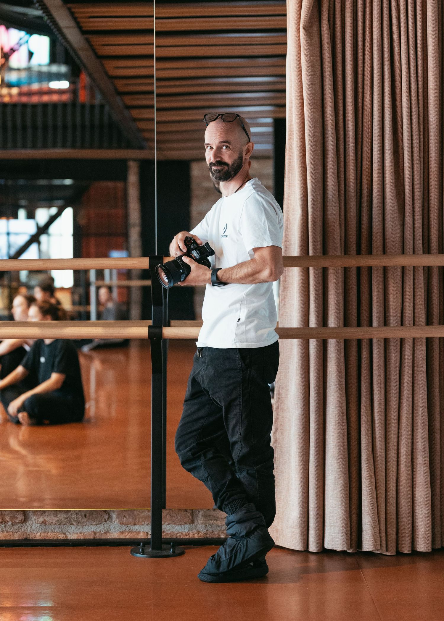 A bearded photographer holding a camera leans against a pole in a dance studio with wooden floors.